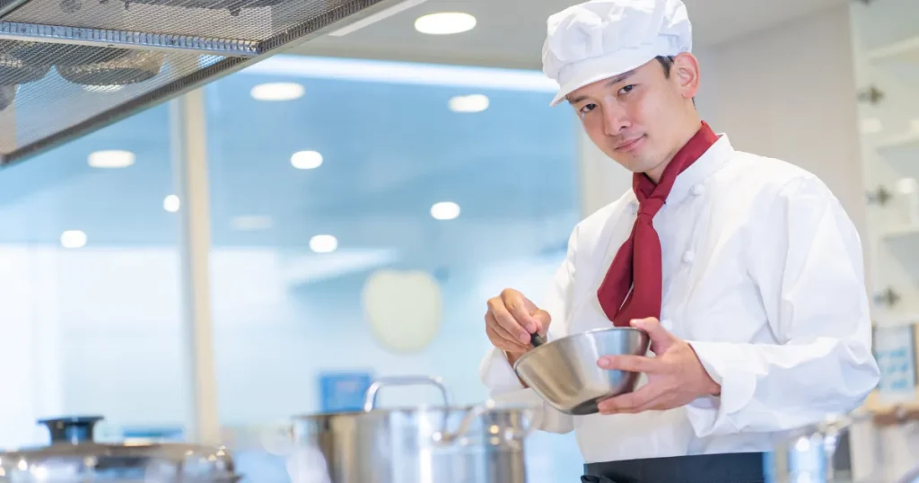 白いシェフ帽とコートを着た料理人がキッチンで調理中。ボウルを手に持ち、調理の基本を実践している様子。料理人の仕事の現場を表現。