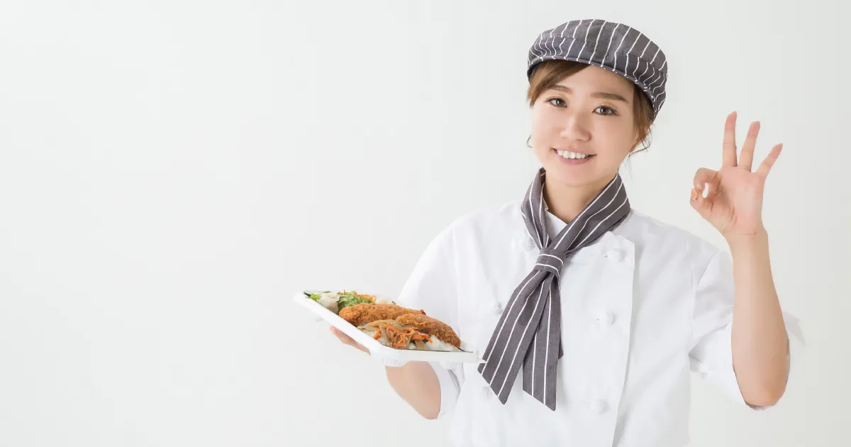 笑顔でお弁当を持ちOKサインを示す惣菜スタッフ。清潔感のある制服と接客姿勢。