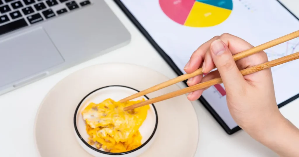 パソコンとグラフ資料が置かれたデスクで、惣菜を箸でつまむ様子。惣菜の試食イメージした構図。