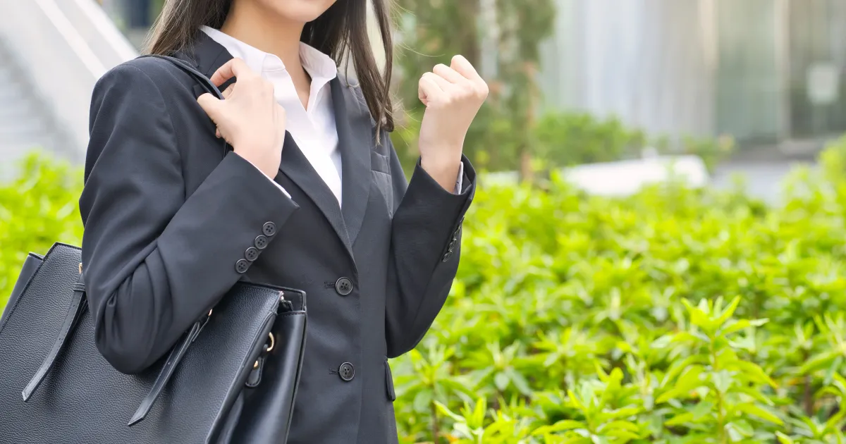 スーツを着た女性がバッグを肩にかけ、片手を握りしめてガッツポーズをしている。背景は緑が広がる屋外の光景で、前向きな姿勢と決意を象徴している。