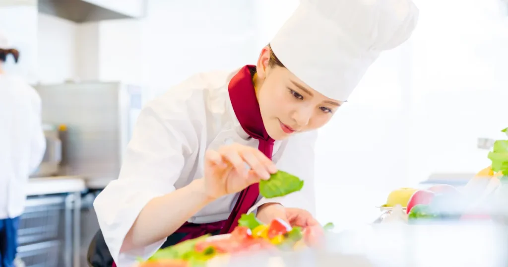 コック帽と調理服を着た女性調理師が、料理の盛り付けに細心の注意を払いながら作業している様子。