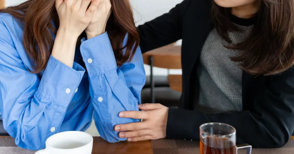 青いシャツを着た女性が顔を手で覆い、悩んでいる様子。その隣に座る人物が優しく手を添えて寄り添い、サポートしている場面。