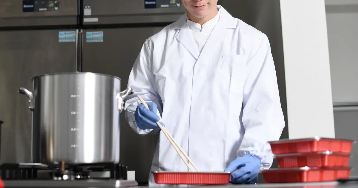 白衣と青い手袋を着用した惣菜スタッフが、調理済みの食品を容器に詰めている様子。衛生管理が徹底された作業環境。