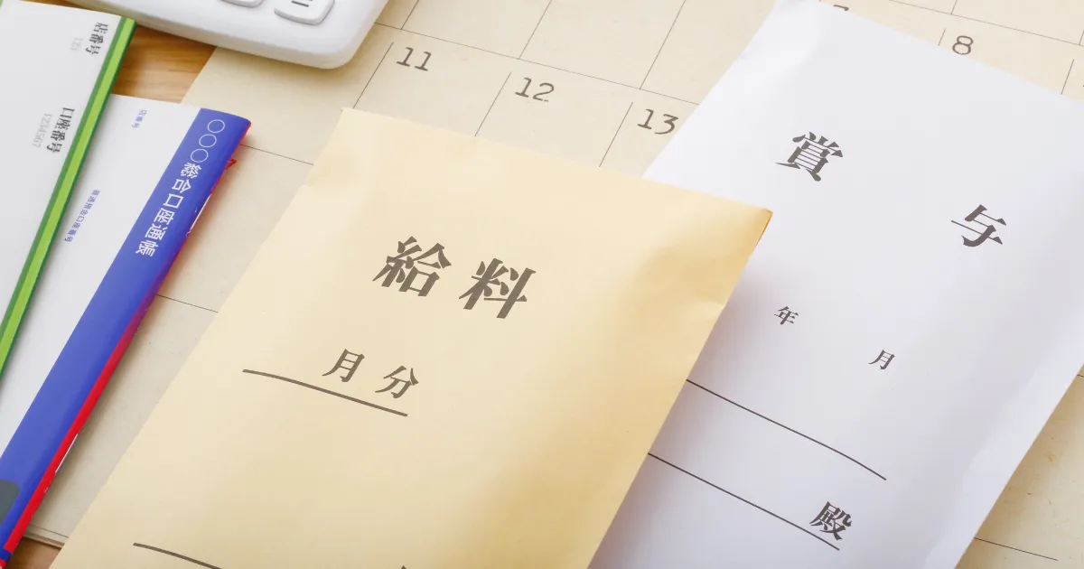給与明細と賞与の封筒、カレンダー、電卓が並ぶデスクの上の様子。調理師・料理人の給料と待遇について説明するイメージ。