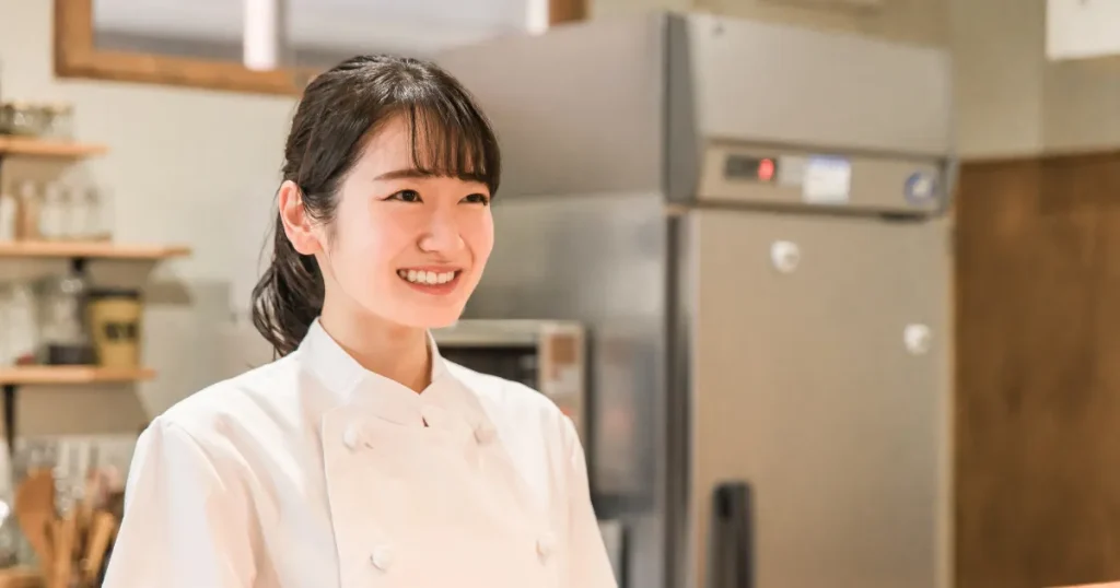 白いシェフコートを着た女性料理人が笑顔で立つ様子。調理師・料理人としてのやりがいや魅力を象徴するイメージ。