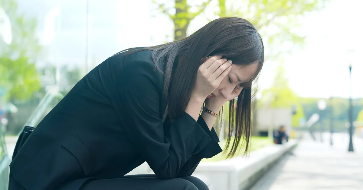 スーツを着た女性が屋外でベンチに座り、頭を抱えて悩んでいる様子。就職活動中のストレスやプレッシャーを象徴する場面。
