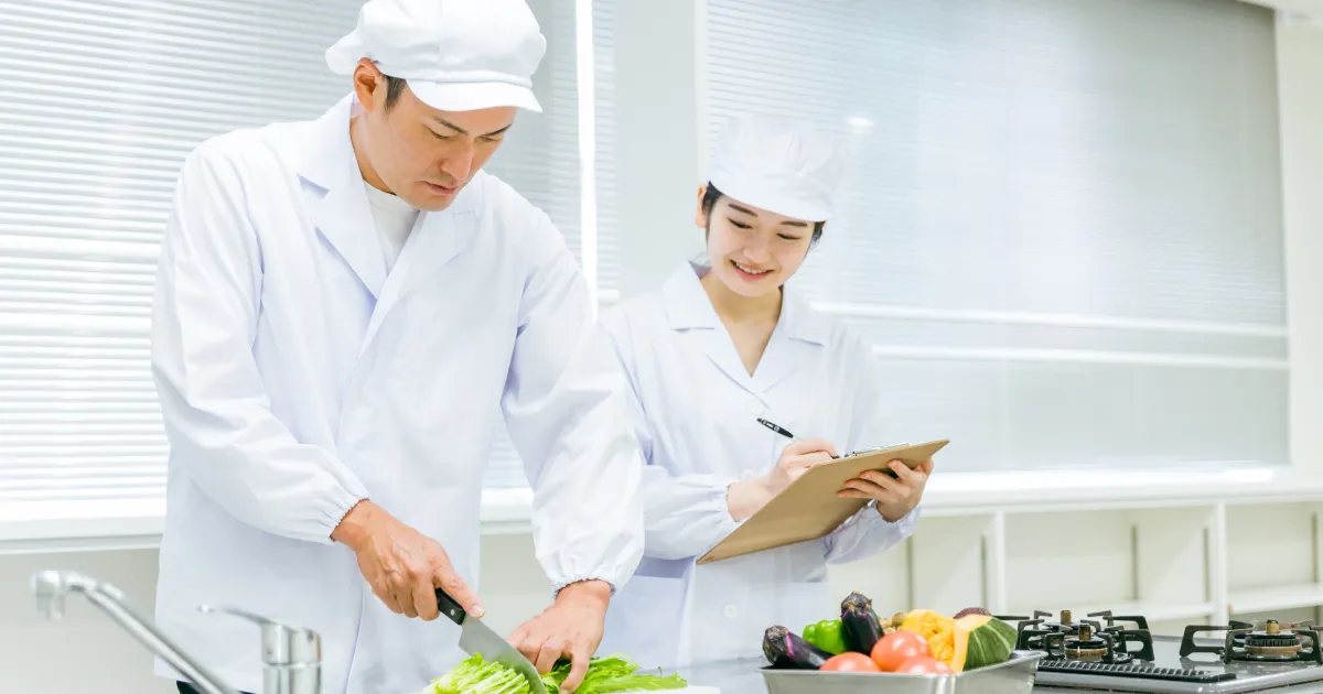 清潔な白衣を着用した調理スタッフが、野菜をカットしながら作業を進める様子。隣ではスタッフがチェックリストに記入している。
