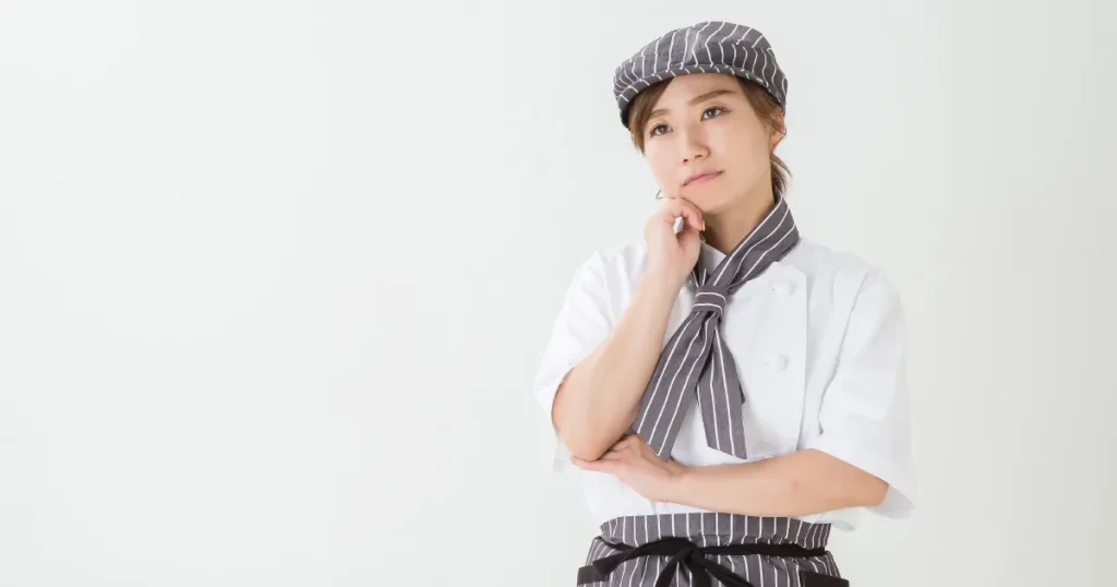 ストライプ柄のエプロンと帽子を着用した食品加工スタッフが、考え込むような表情で腕を組んでいる姿。