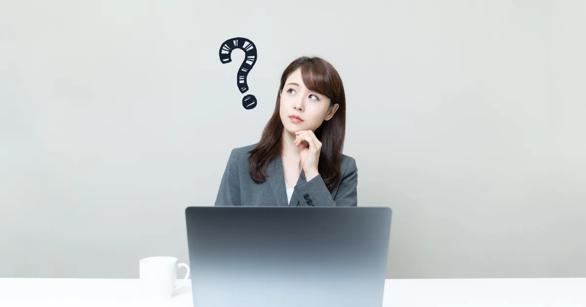 スーツを着た女性がパソコンの前で考え込んでいる様子。頭上にクエスチョンマークが描かれており、自分の強み・弱みがわからず悩んでいる状況を表現したイメージ。