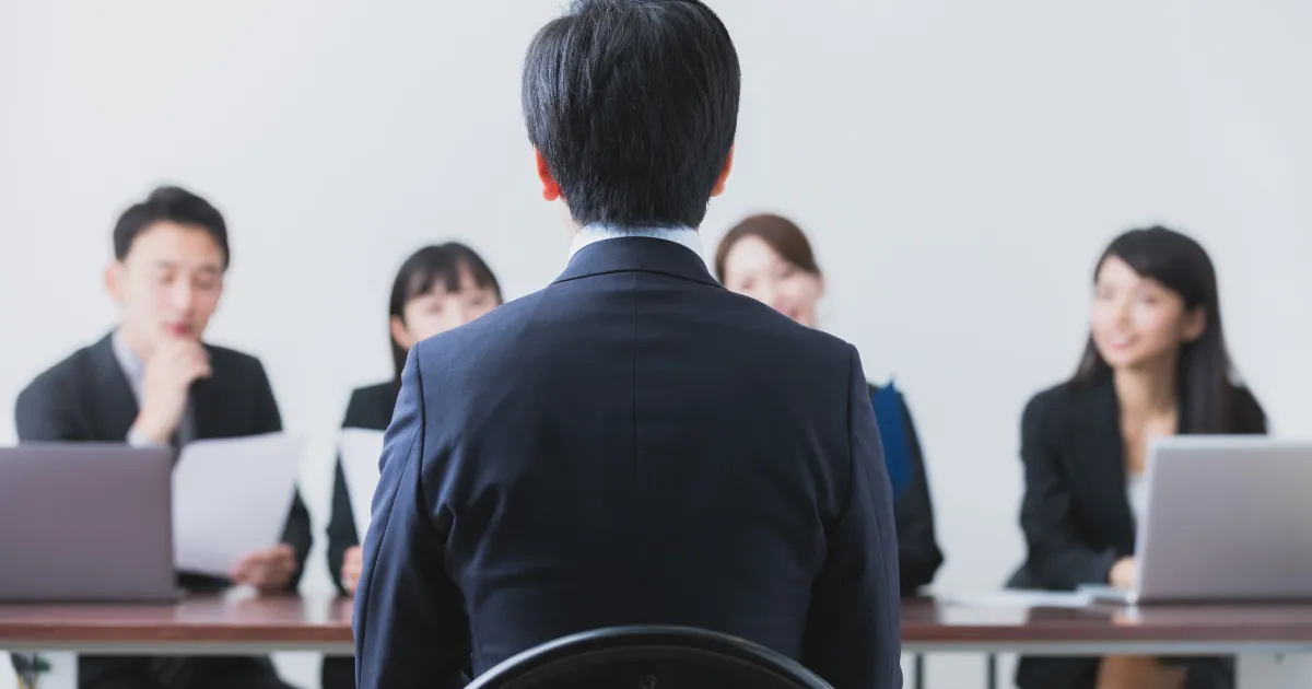 スーツ姿の男性が採用面接で面接官たちと向き合っている様子。企業が就活生に『自分の強み』を尋ねる理由を説明する場面をイメージした画像。