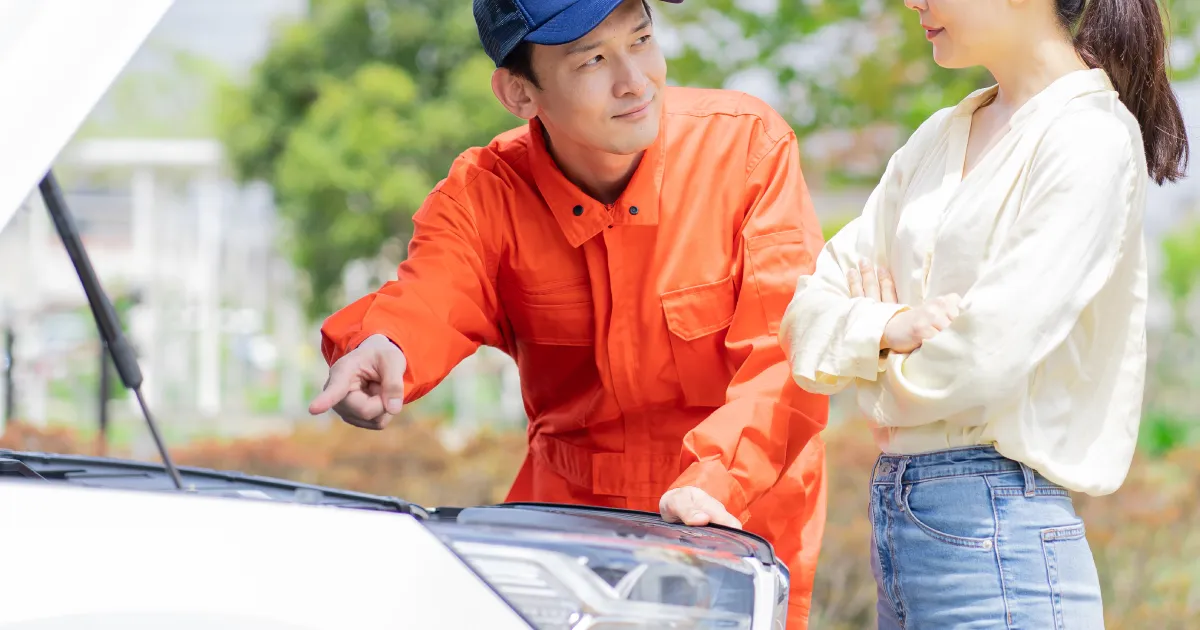 オレンジ色の作業服を着た自動車整備士がボンネットを開けて車の状態を指し示しながら、顧客と話している場面。