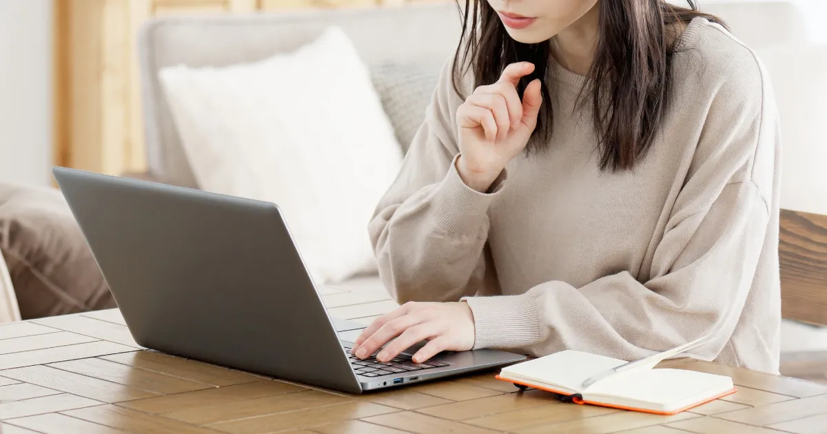 ノートパソコンを操作しながら転職先を探している女性。横にノートが置かれた木製テーブルが映る。