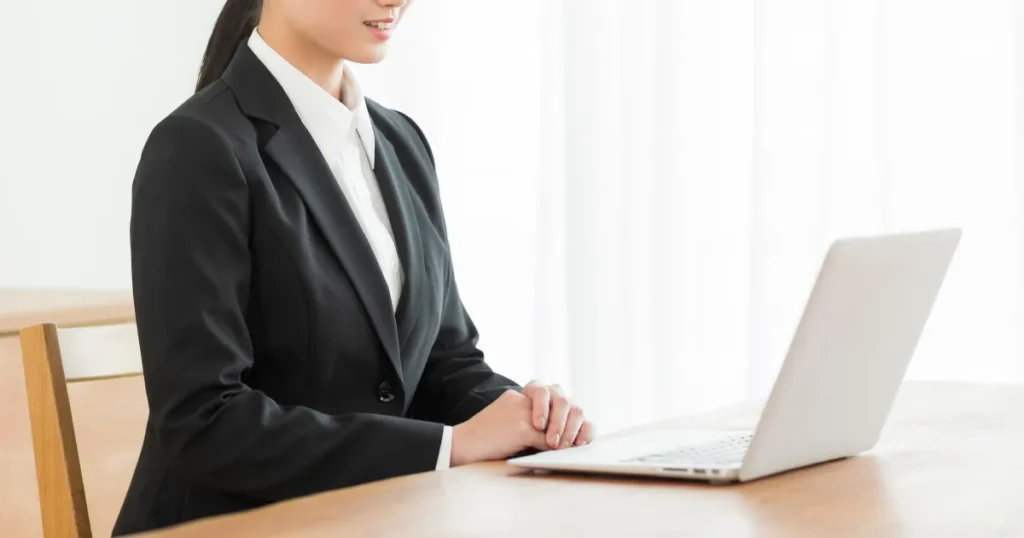 Web面接の準備を整え、ノートパソコンの前で姿勢よく座るスーツ姿の女性。オンライン面接前の確認を行う様子。