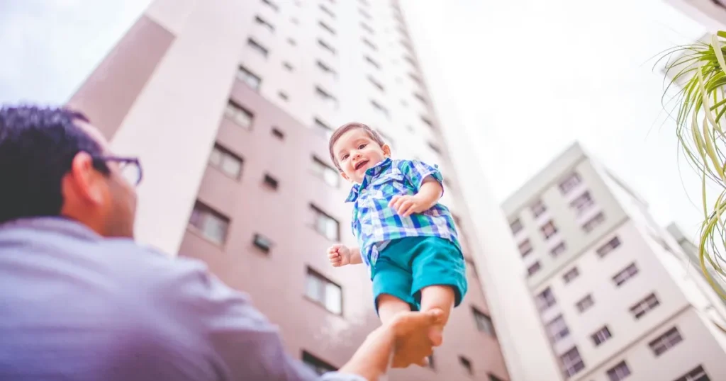 高層マンションの前で赤ちゃんを抱き上げる父親。育休中の転職をスムーズに進めるためのステップを表現したイメージ。