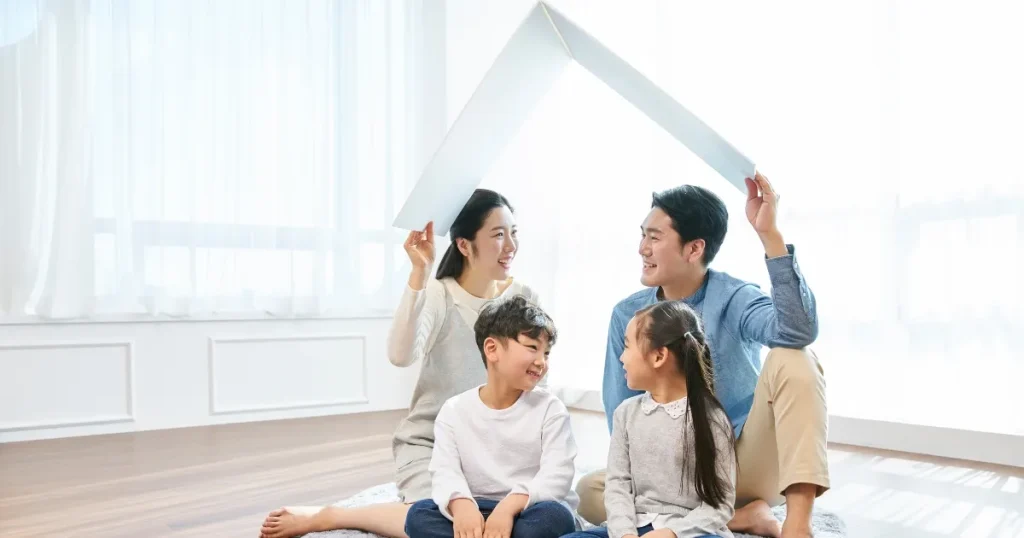 リビングで家族4人が集まり、両親が紙で作った屋根を子どもたちの上にかざしている様子。安心感や保護を象徴するイメージ。