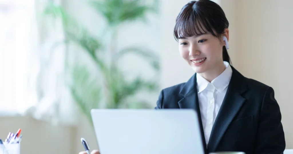 Web面接中に笑顔でパソコン画面を見つめる女性 - 清潔感のある髪型でスーツを着用し、イヤホンを装着