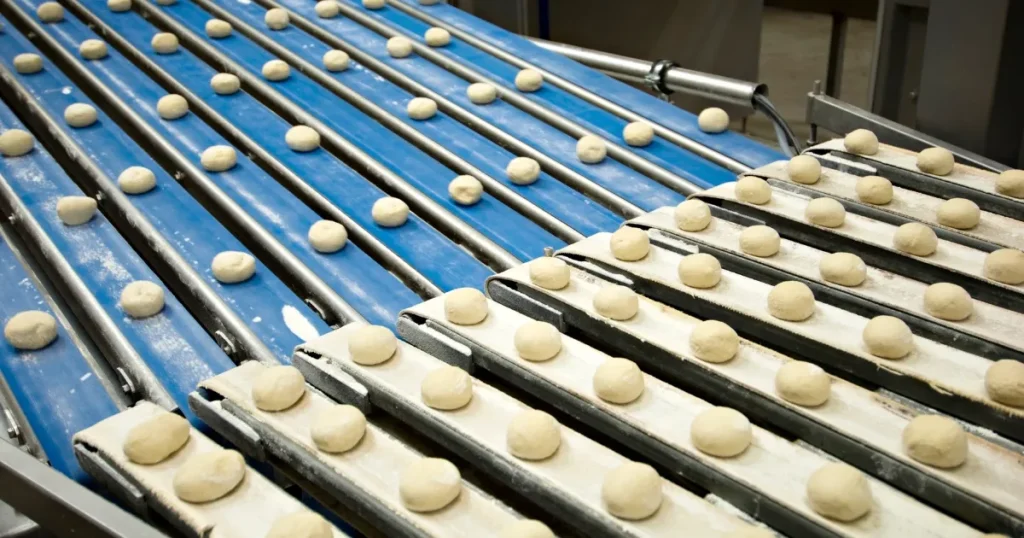 食品メーカーの製造ラインで均一に並べられたパン生地が、ベルトコンベアで運ばれている様子。効率的な生産と品質管理を象徴するシーン。