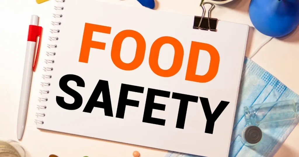 『FOOD SAFETY』と大きく書かれたメモ帳がテーブルに置かれており、隣にはペン、マスク、試験器具が配置されている。食品安全を象徴するイメージ。