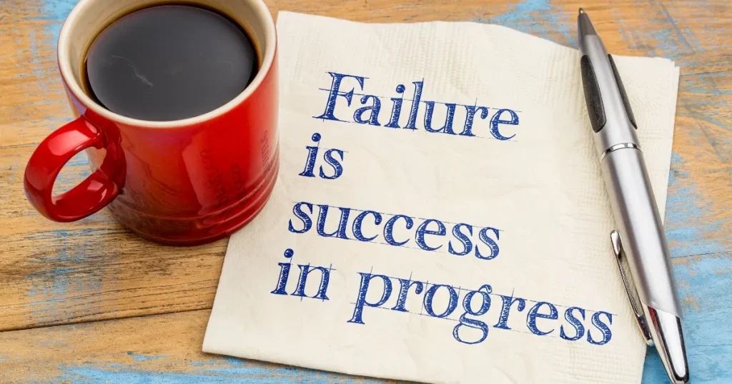 木製テーブルに置かれた赤いコーヒーカップとペン。ナプキンには『Failure is success in progress』と書かれており、失敗を成功へのプロセスとして捉えるメッセージを伝えている。
