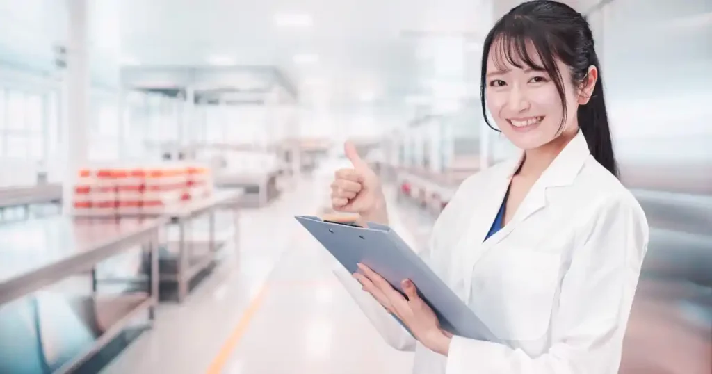 食品メーカーの工場内で白衣を着た女性スタッフがクリップボードを持ち、親指を立てて笑顔でポーズを取っている。食品業界で働く適性や積極性を象徴するイメージ。