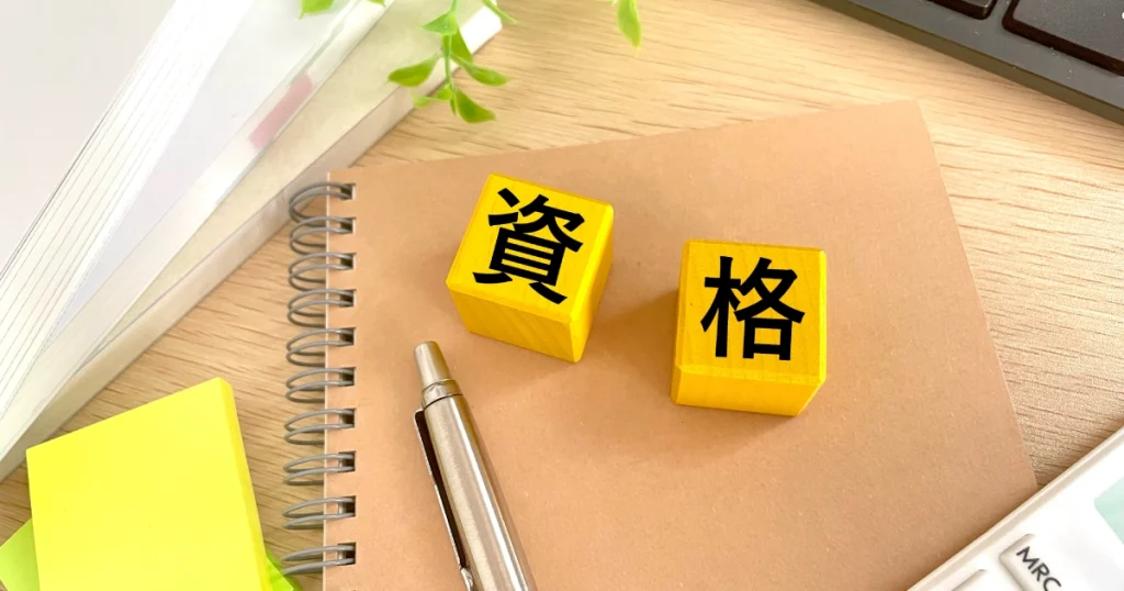 資格の文字が書かれた黄色のブロックとノート、ペンが置かれたデスク - 転職に役立つ資格の選び方を説明するセクション用の画像