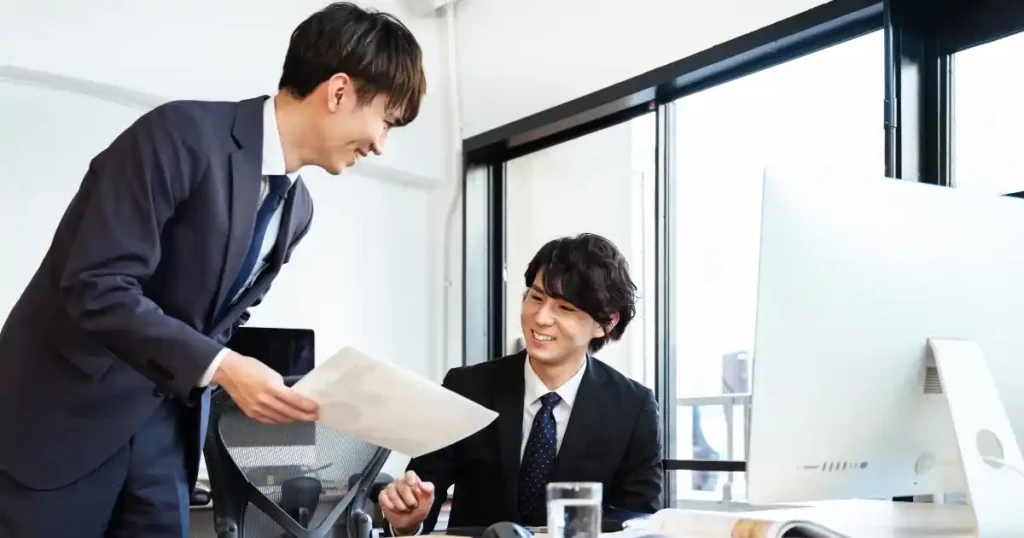オフィスで笑顔で話し合うスーツ姿のビジネスパーソン - 年収を上げるための具体的な方法を説明するセクション用の画像