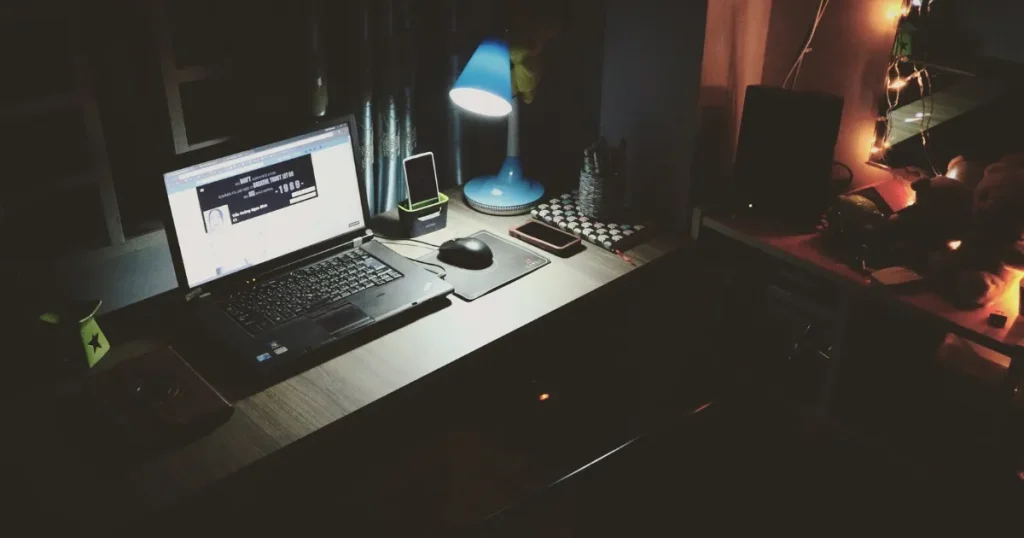 暗い部屋のデスクに置かれたノートパソコンとスタンドライト。Webテストが企業における採用活動でどのように活用されるかを象徴するイメージ。