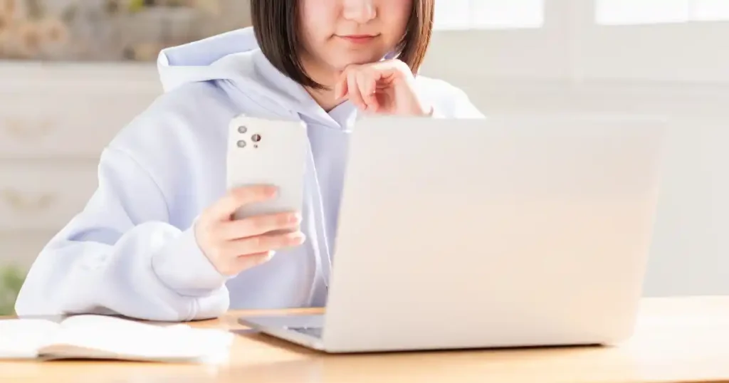 パソコンとスマートフォンを使いながら就職活動の調べ物をしているフード付きパーカーを着た女性。