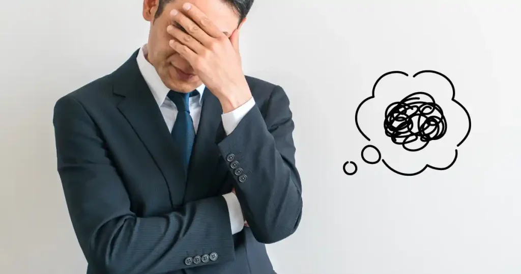 スーツを着た男性が頭を抱え、隣に悩んでいる思考を表す吹き出しが描かれているイメージ。
