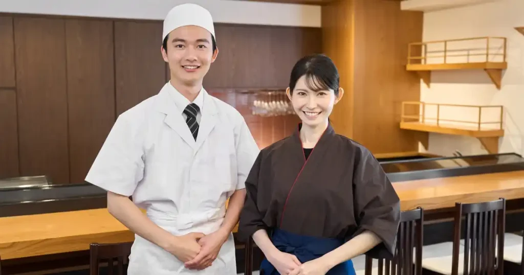 寿司職人とスタッフが調理場で並んで笑顔を見せる。清潔感とプロフェッショナルを感じさせる服装を着用。