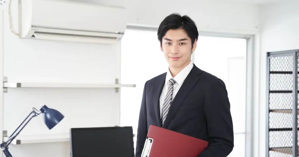 スーツを着た若い男性がファイルを持って笑顔で立つオフィスの風景 - インターンシップに参加するメリットを表現するイメージ