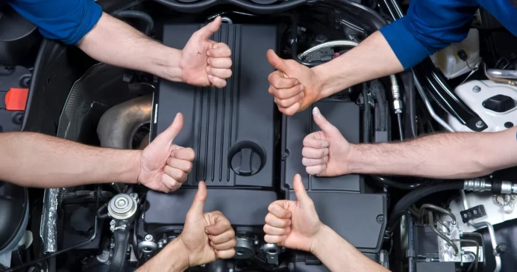 自動車エンジンルームの上で親指を立てて成功を祝う複数の手。自動車検査員資格取得のプロセスを象徴するイメージ。