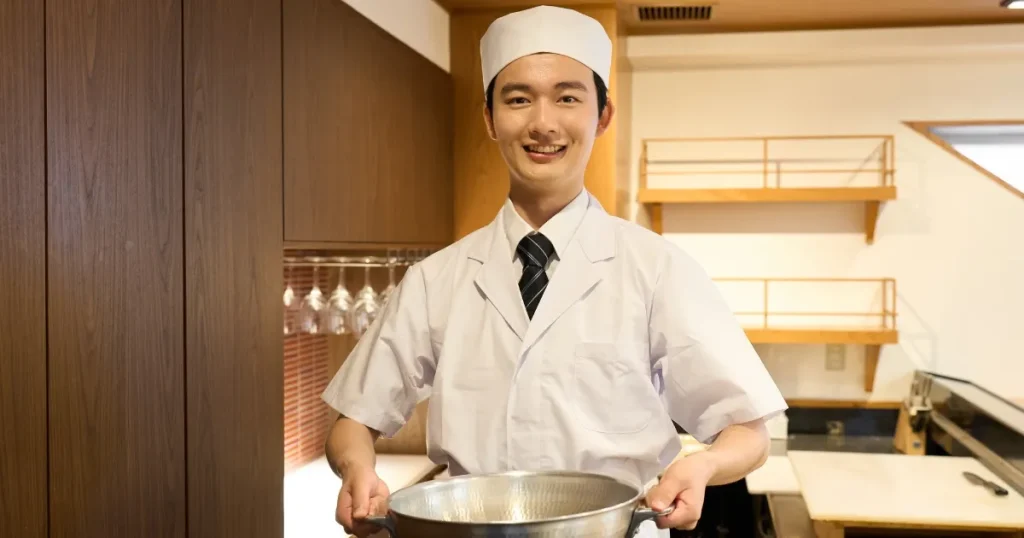 笑顔で調理器具を持つ寿司職人。清潔な白衣を着用し、和やかな雰囲気の店内で撮影されたシーン。