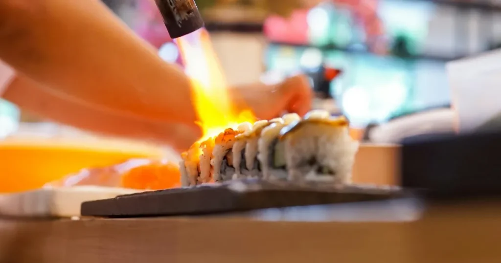 寿司職人がバーナーを使用して寿司を調理しているシーン。焼き目がつけられた寿司が並ぶ躍動感あふれる光景。