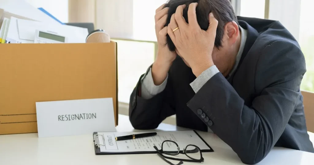頭を抱えるスーツ姿の男性と、デスクの上に置かれた『RESIGNATION』と書かれた紙や書類。離職率が高い業界からの転職における課題を象徴するシーン。