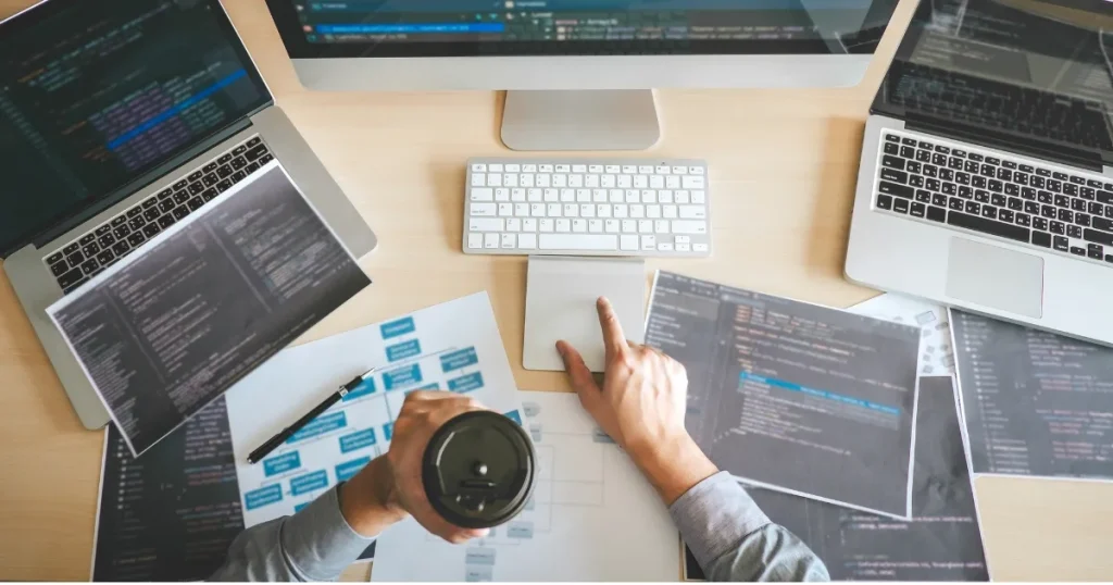 複数のモニターとコードが記載された資料を見ながら作業を進める人の手元。IT業界での文系出身者の活躍を象徴するシーン。
