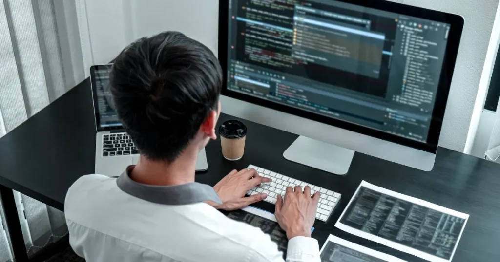 プログラミングコードが表示されたモニターの前で作業するエンジニア。IT業界の仕事内容を象徴するシーン。
