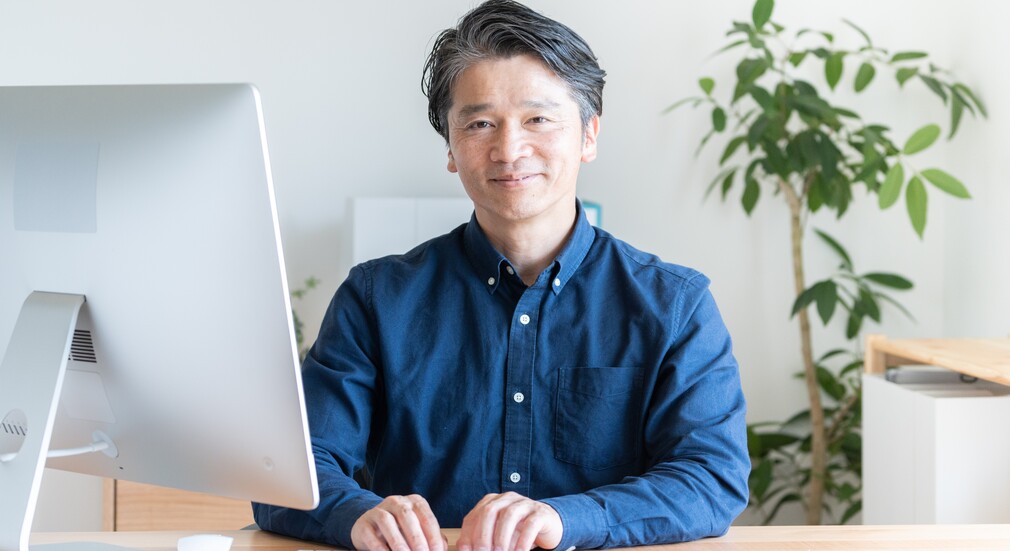 パソコンの前で微笑むカジュアルなシャツを着た男性。50代転職の注意点を象徴するイメージ。