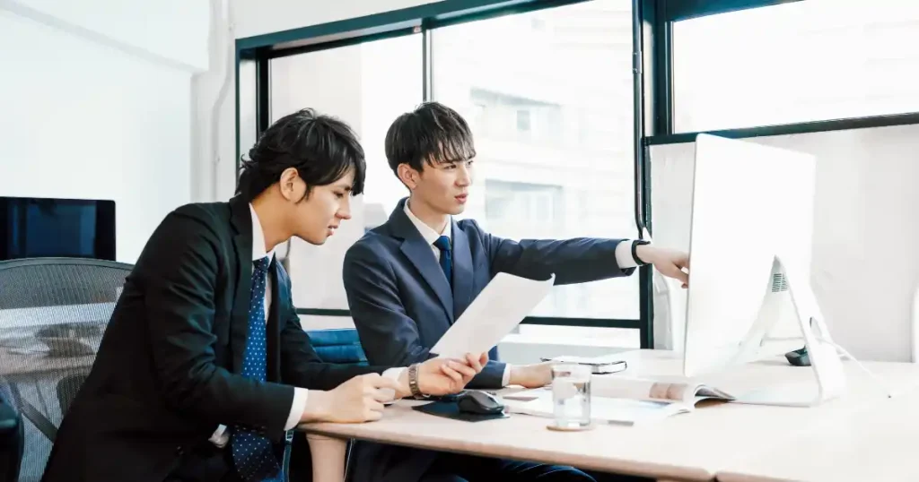 オフィスでディスプレイを見ながら資料を確認する2人のビジネスマン。社会人インターン先を選ぶ際の職場環境や指導体制を検討する場面を象徴している。