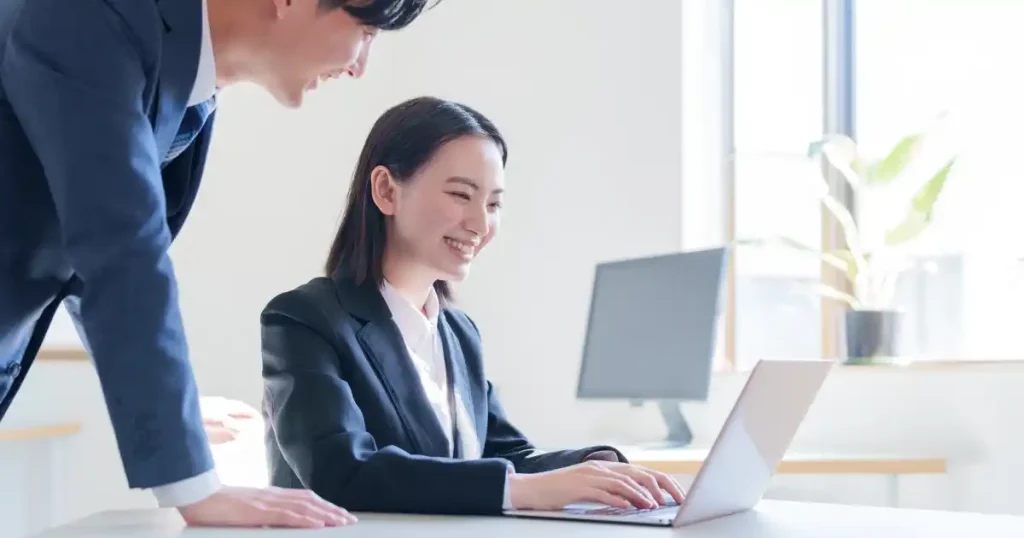 ノートパソコンで作業を進める女性と、それをサポートする男性。同僚との協力やフィードバックを通じて社会人インターンを成功させるポイントを表現している。