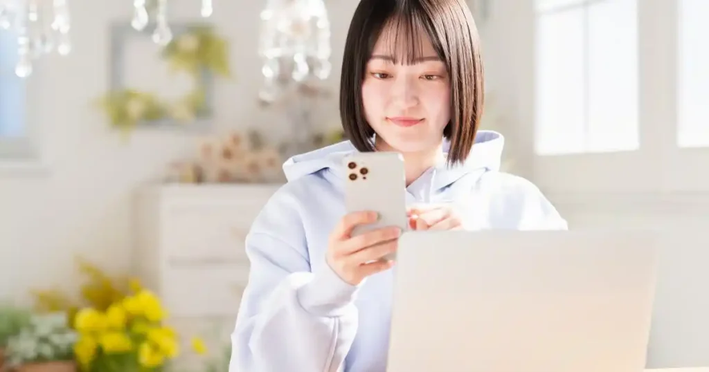 スマートフォンを操作しながらノートパソコンに向かう女性：企業情報を検索し、就活での企業選びを進めている様子を表現。