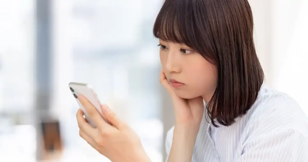 スマートフォンを見つめながら悩んでいる様子の女性。就職活動中の不安や悩みを抱えている姿を表現したイメージ。