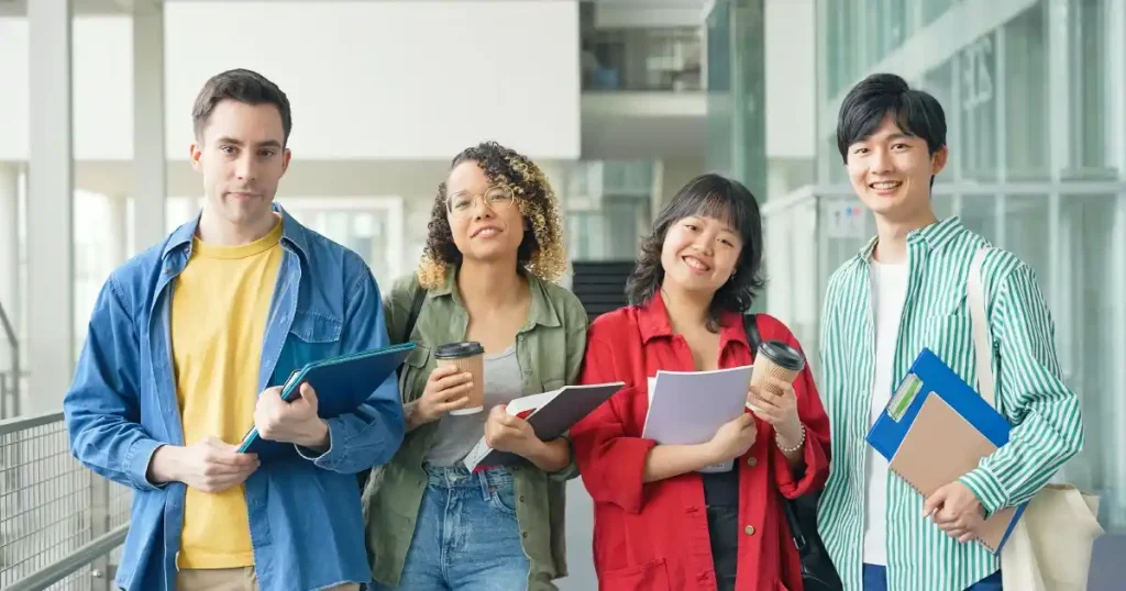 大学のキャンパス内で立ち話をしている多国籍の学生たち。ノートやファイルを手に持ちながら、笑顔で親しげな様子を見せている。