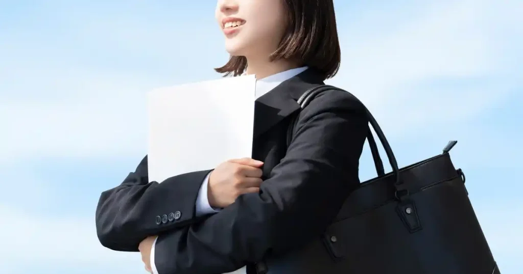 空を背景にスーツを着た女性が資料を抱え、明るい表情で前を見つめている。これから就活を始める希望に満ちた姿を象徴するイメージ。