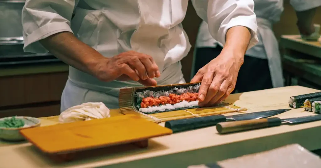 寿司職人が巻きすを使って細やかに巻寿司を仕上げている様子。カウンターには包丁や調理道具が並べられ、職人の丁寧な手仕事が感じられる。