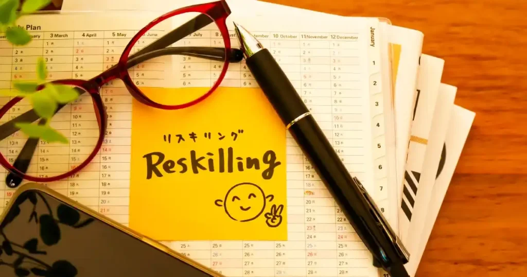 「リスキリング」と書かれたメモ帳、ペン、眼鏡がデスクに置かれている画像。新しいスキルを学び直すリスキリングの概念を象徴。